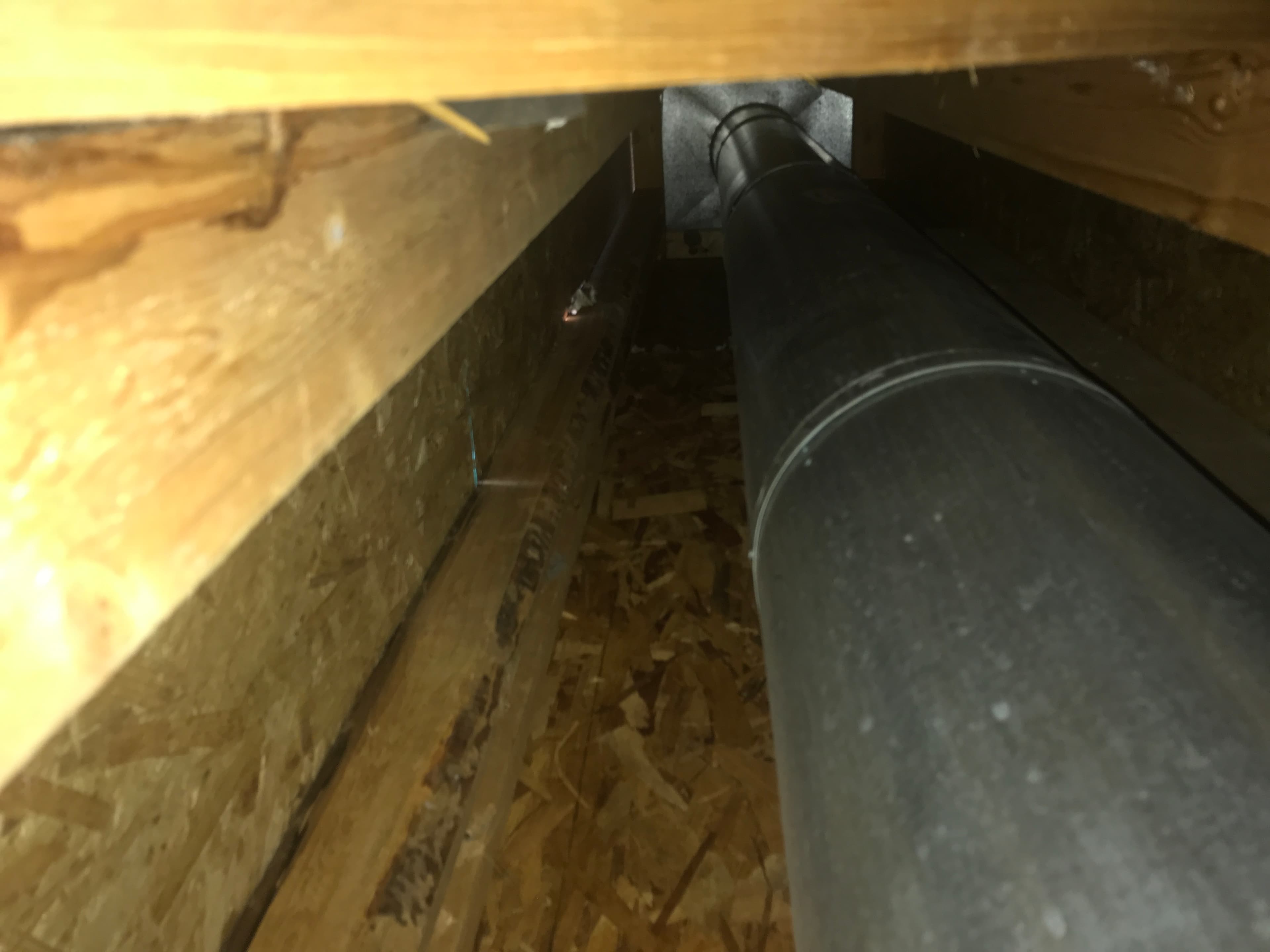 Interior attic inspection showing a metal exhaust vent pipe running along wood rafters and OSB decking.