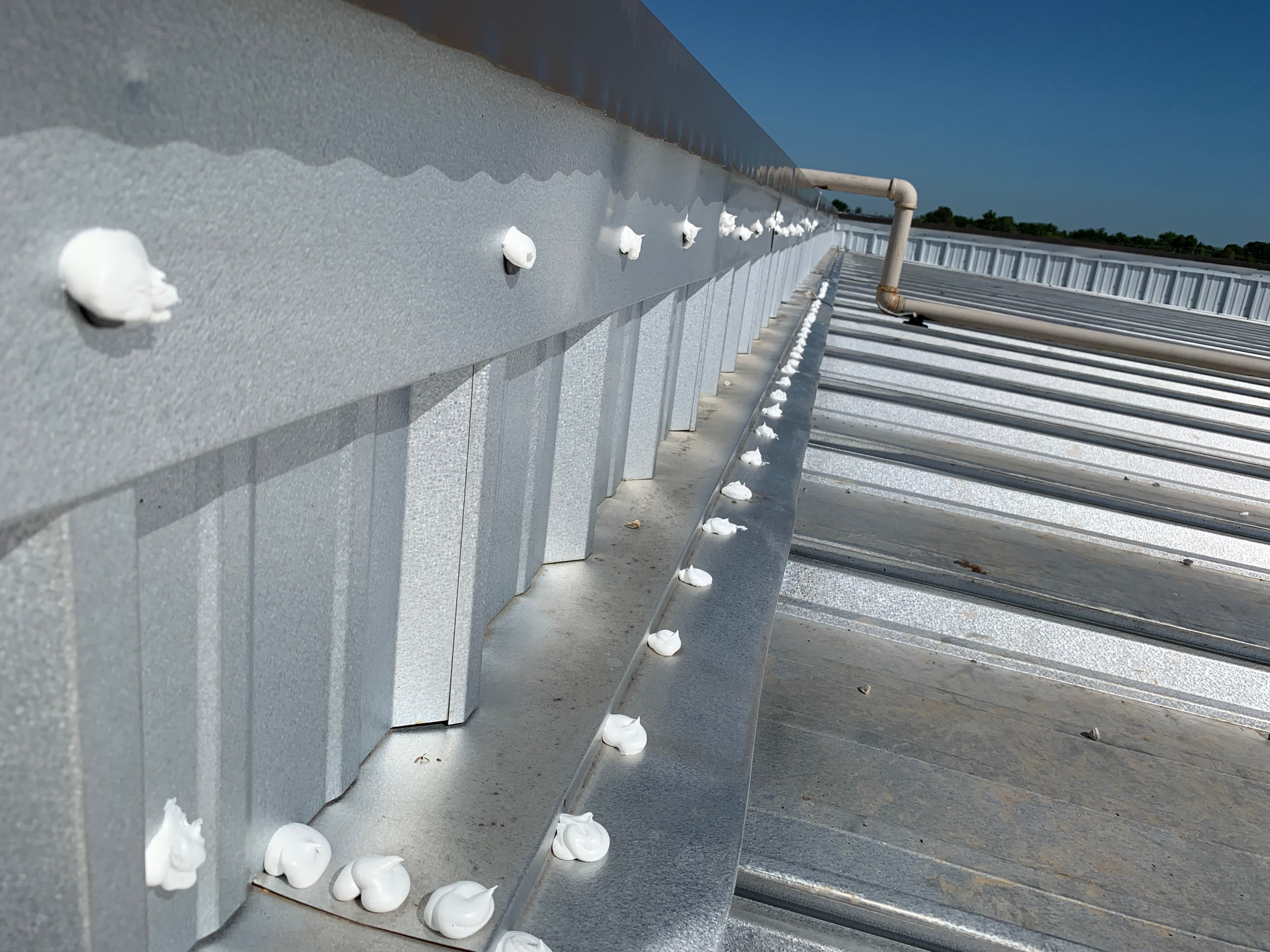 Close-up view of white commercial silicone sealant applied over exposed metal roof panel screws and base flashing.