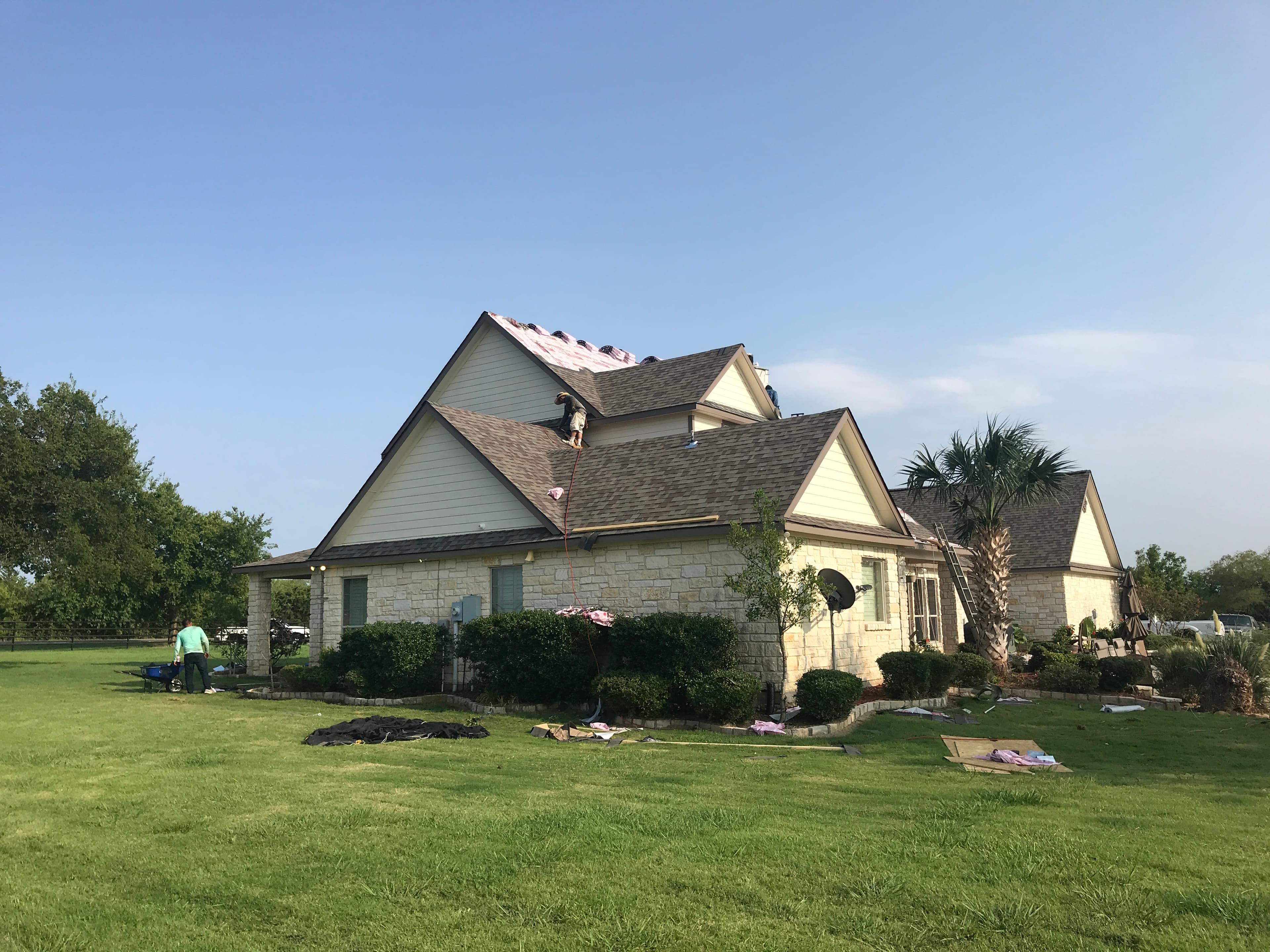 Roofer installing brown architectural asphalt shingles on a multi-gable stone house.