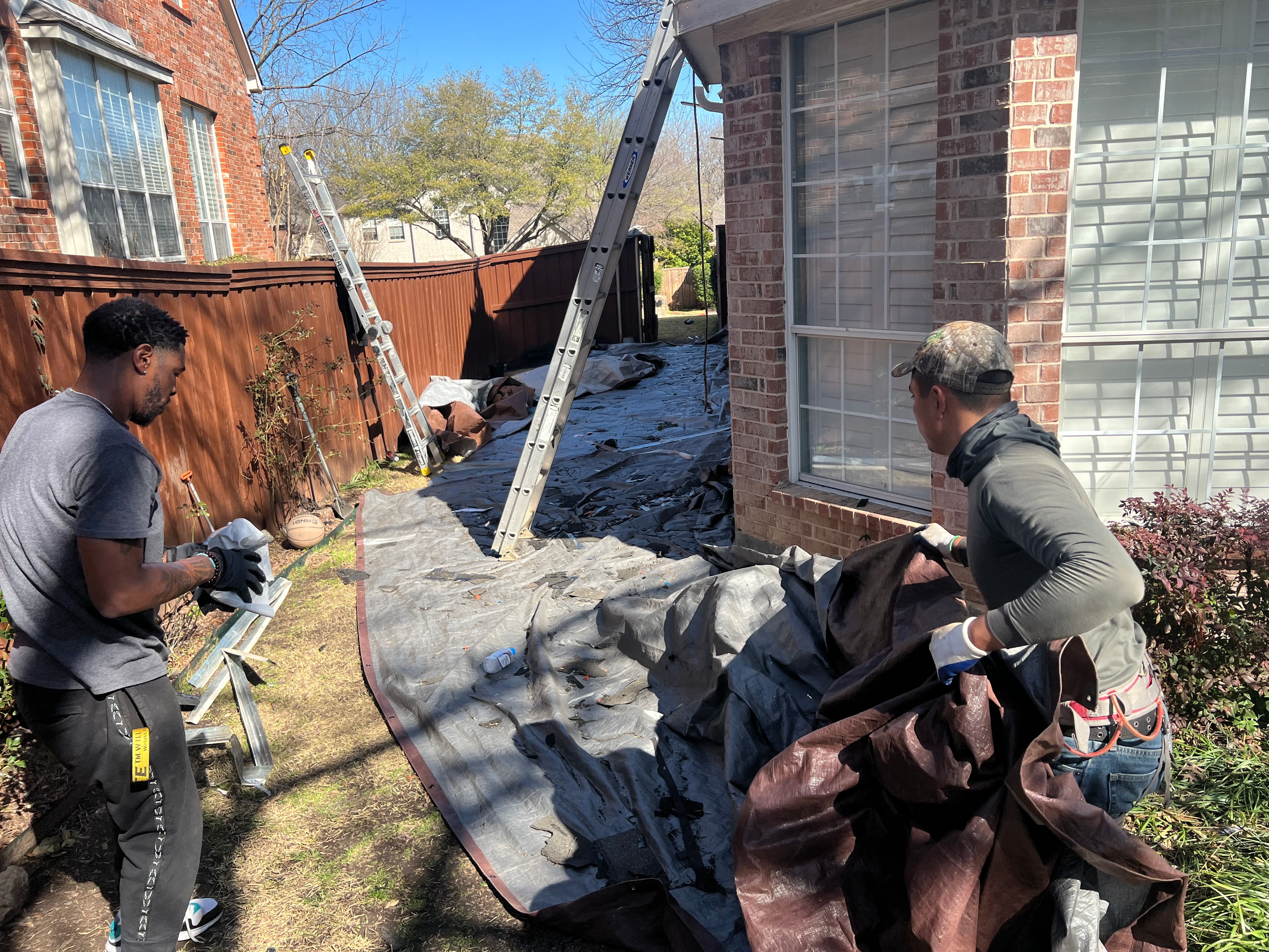 Roofing crew laying down heavy-duty landscape protection tarps along the side yard of a brick home.