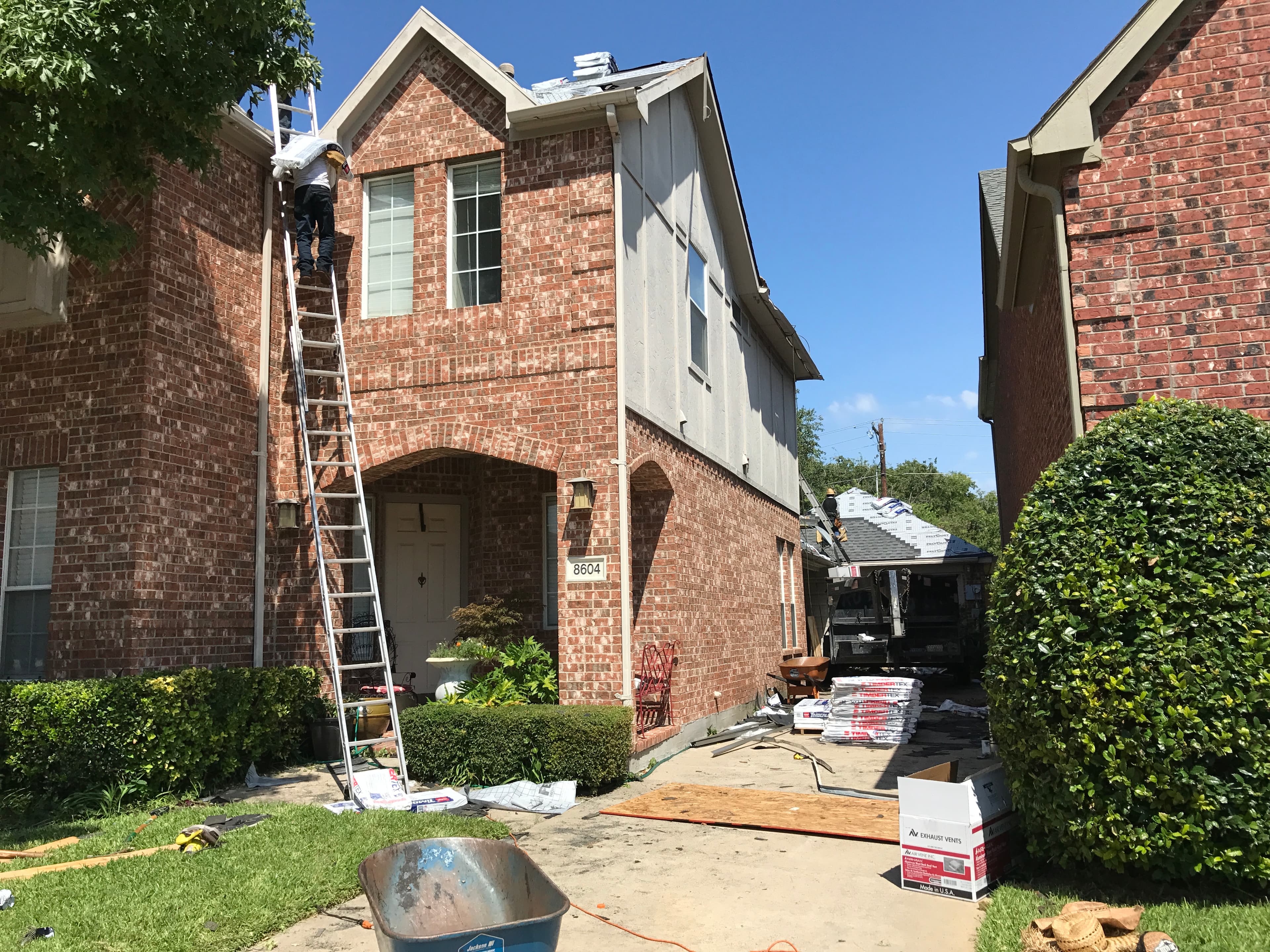 Full view of a residential roofing job site in progress on a clear day, capturing a modern brick house, a prominent tree, and all associated equipment and staged materials.
