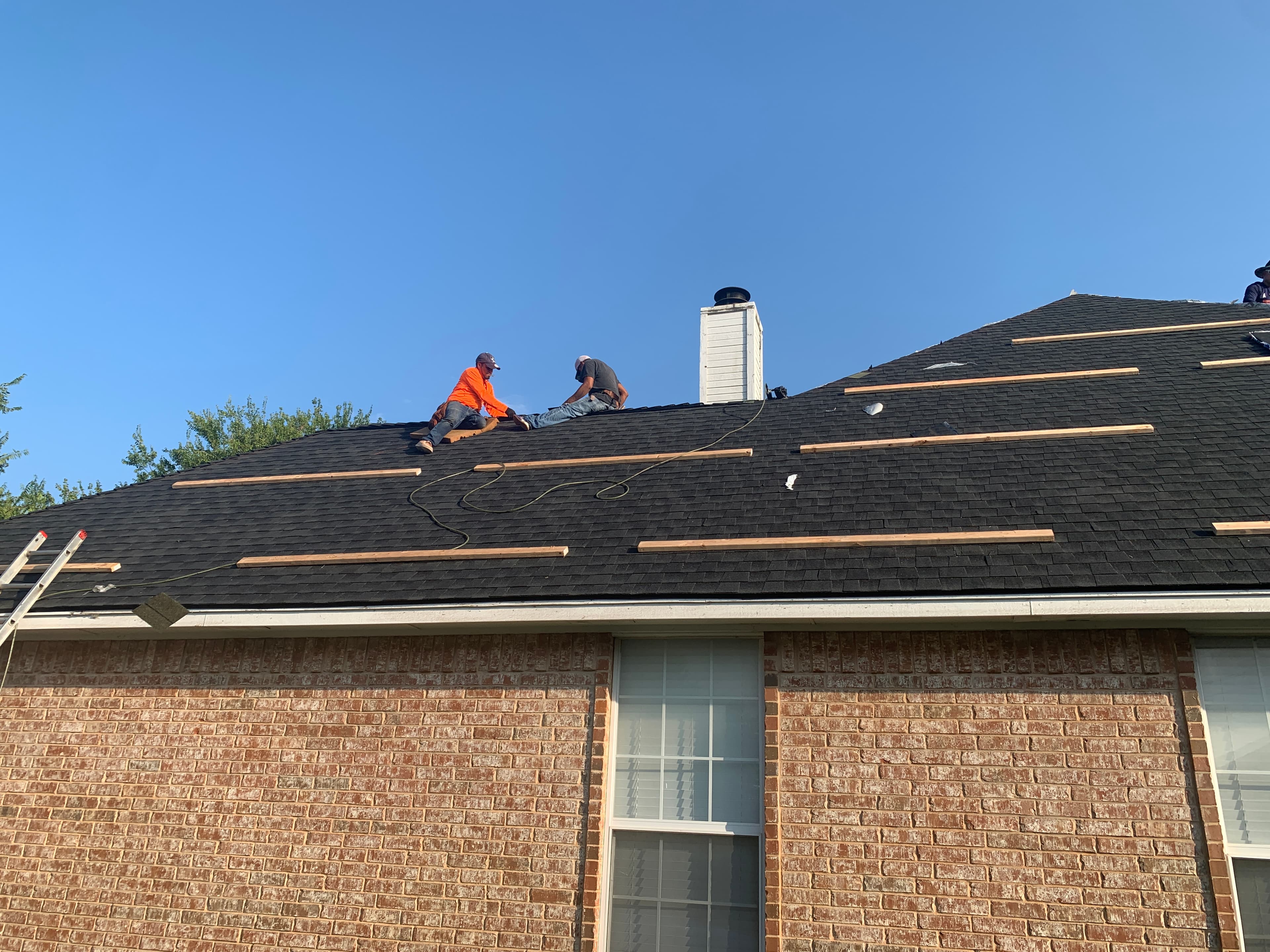 Roofing crew working on a steep-pitch asphalt shingle roof using wooden toe boards for safety near a chimney.