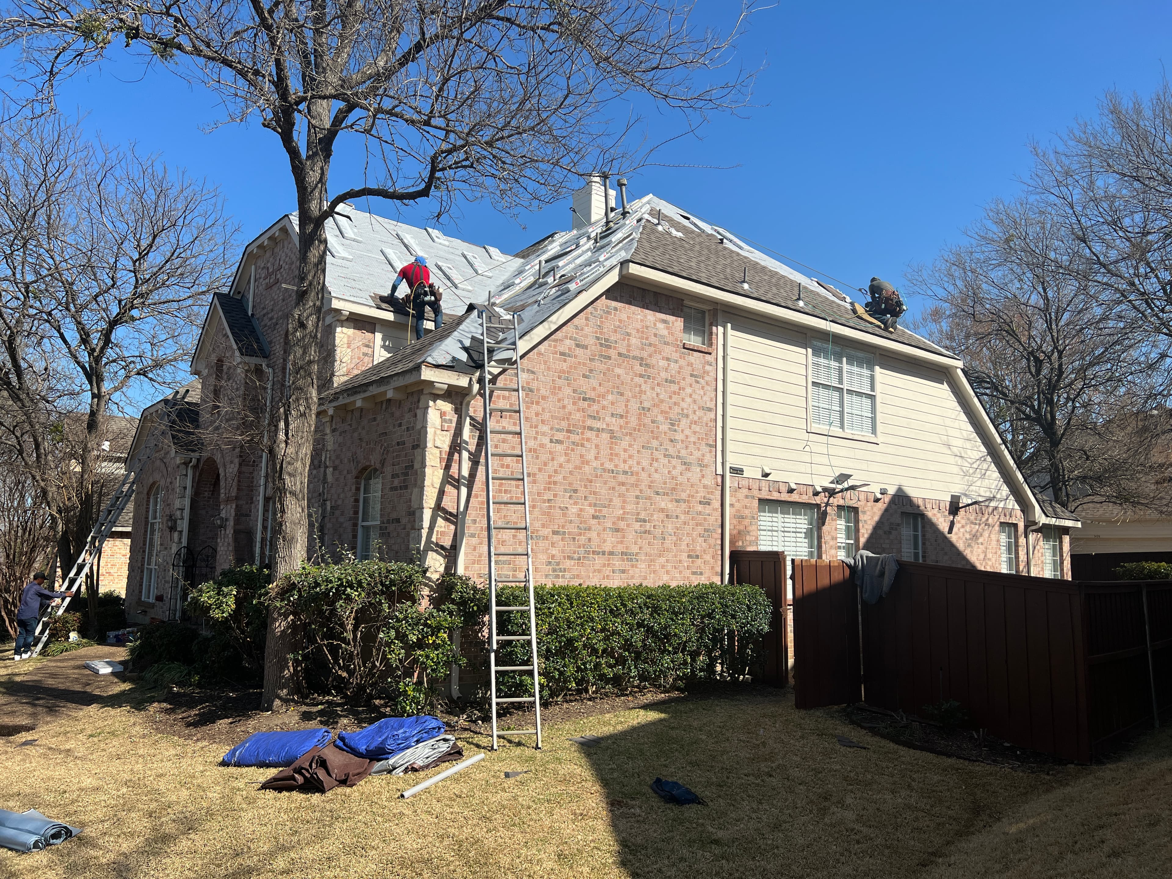 Side elevation of a large brick home showing extension ladders, property protection tarps, and roofers laying synthetic underlayment.
