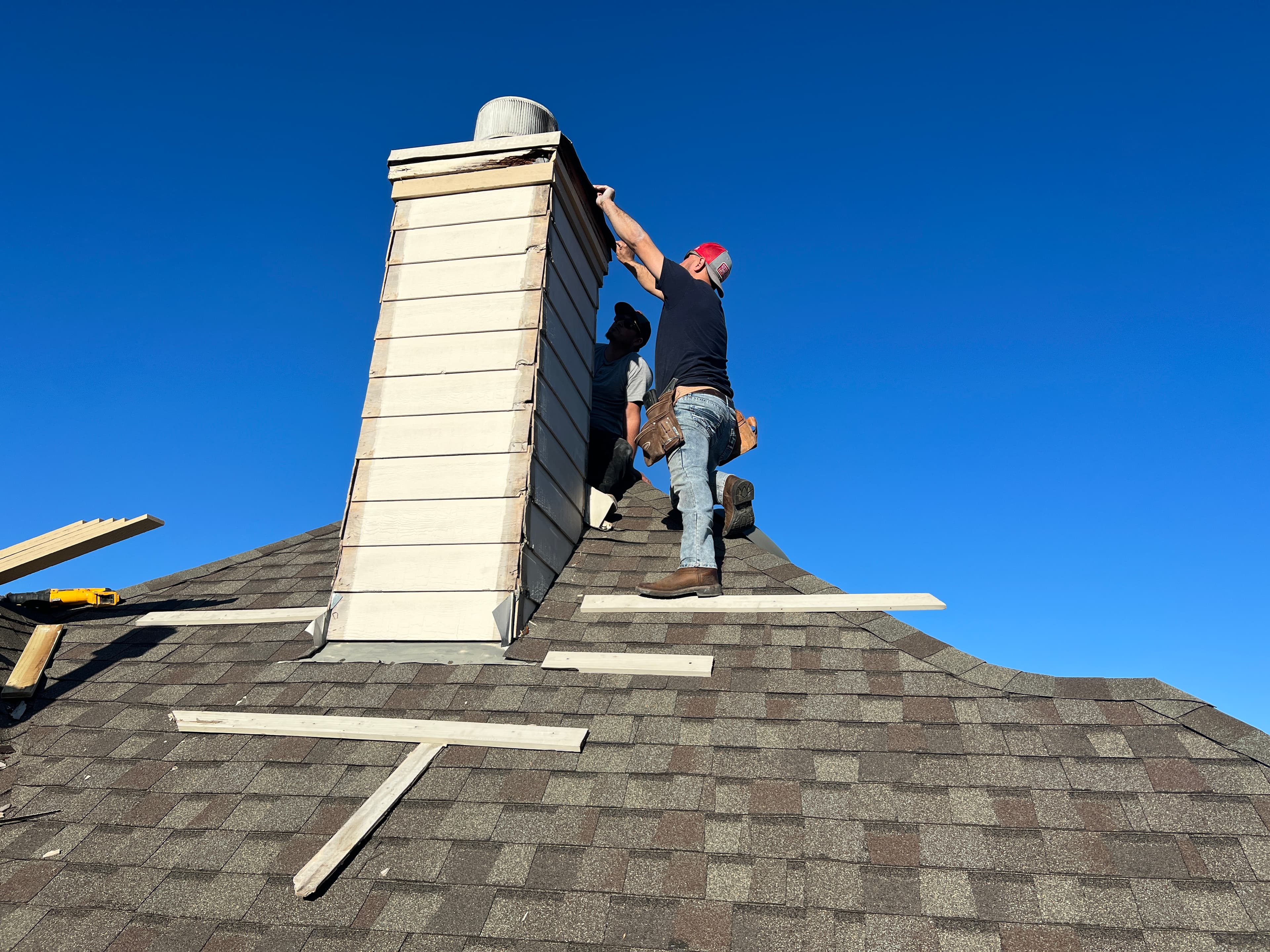 Professional roofers repairing wood siding and flashing around a high-pitch chimney box during a roof replacement.