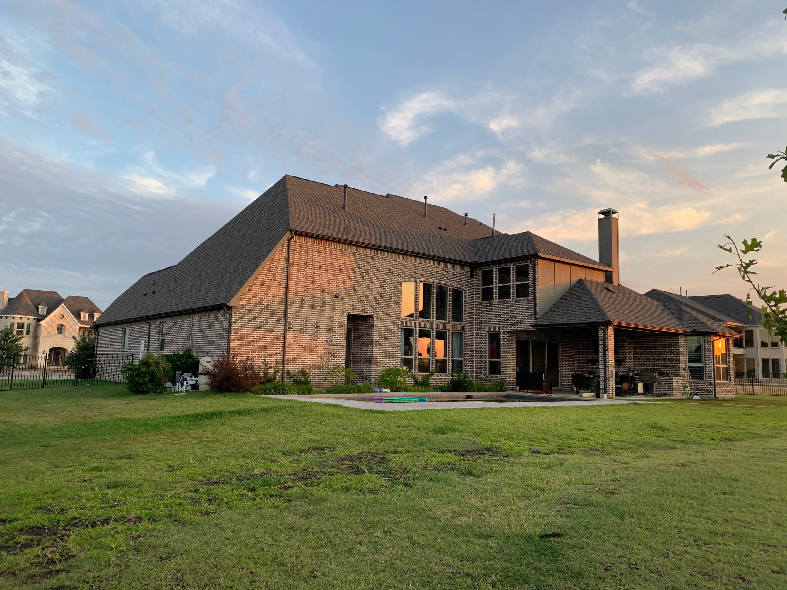 Rear elevation of a custom brick home showcasing a steep-slope architectural shingle roof and covered patio extension near a pool deck.