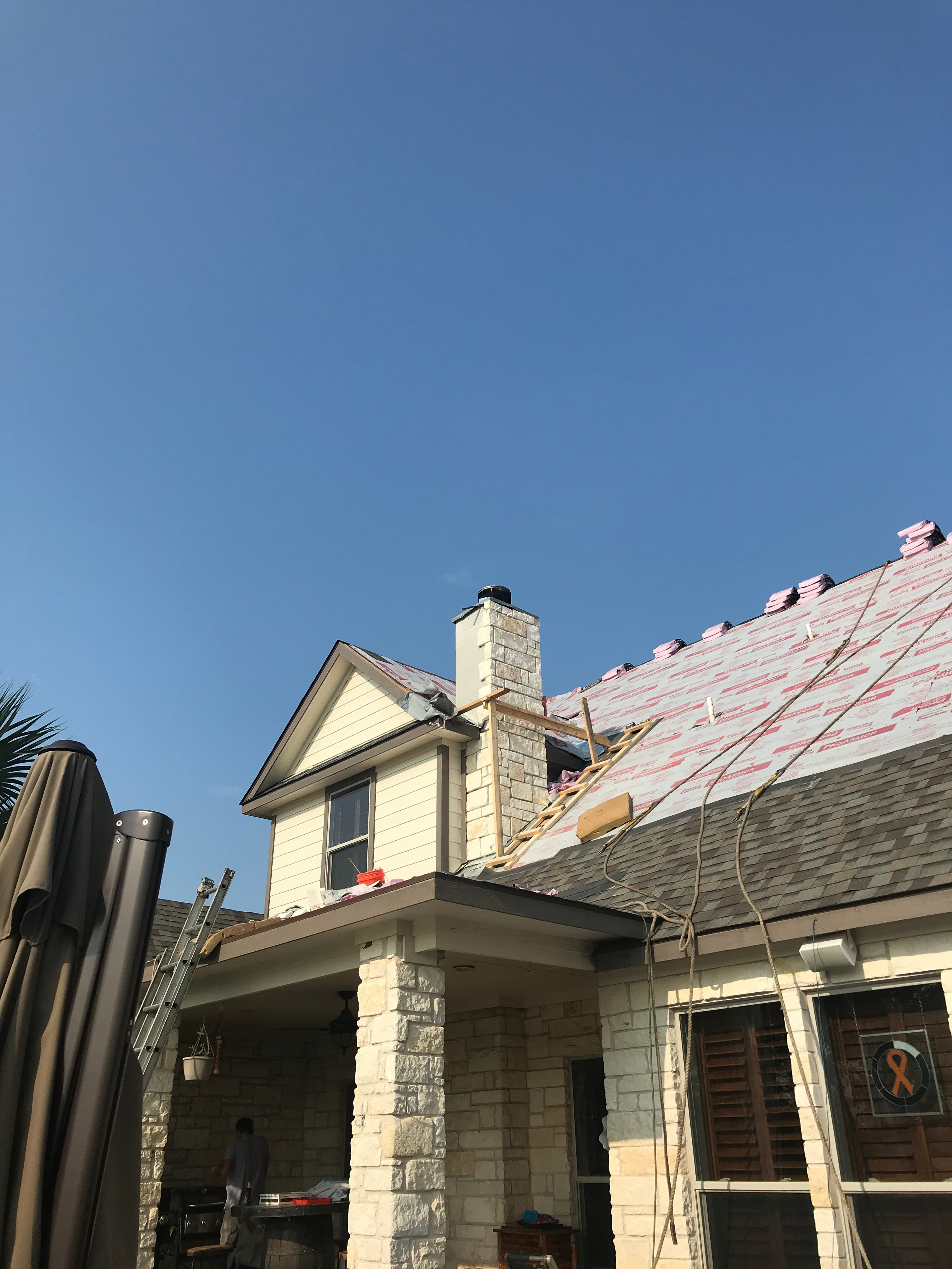 Safety ropes and wooden scaffolding setup around a stone chimney during synthetic roof underlayment installation.