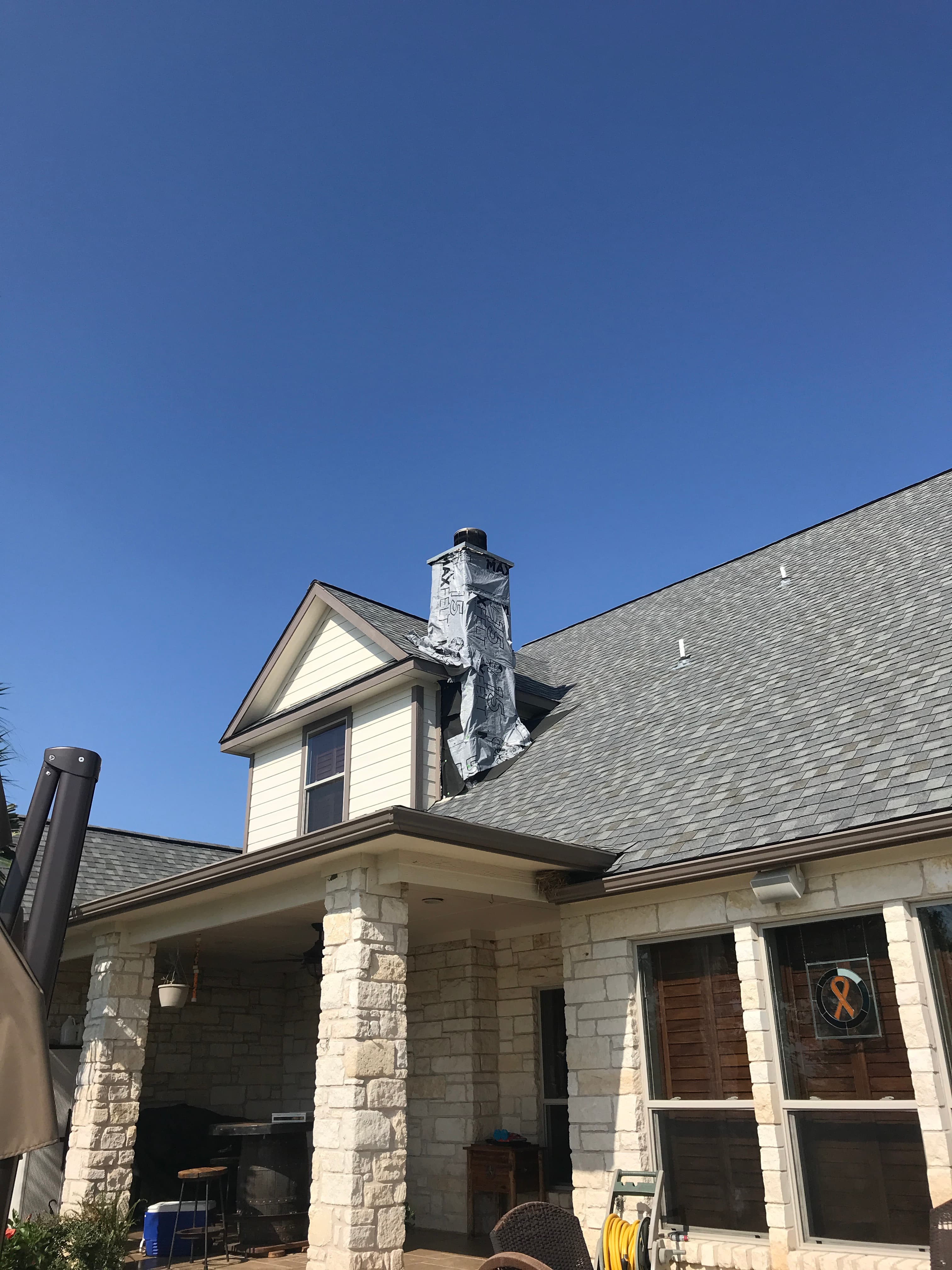 Exterior view of a stone residence with gray asphalt shingle roofing and an emergency weather-wrapped chimney during leak repair.